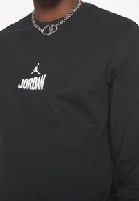 Mustapaita, jossa on pitkät hihat, valkoinen "JORDAN" -logo ja hyppäävän miehen grafiikka; sileää puuvillakangasta, pyöreä pääntie, näkyvissä hopeinen ketjun kaulakoru.