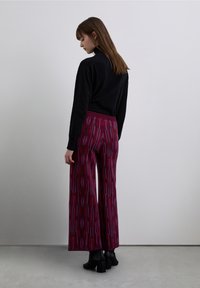 Pantaloni a gamba larga in bordeaux profondo con motivi geometrici; abbinati a un maglione nero e stivaletti neri con tacco a blocco. Tessuto liscio, vestibilità rilassata.