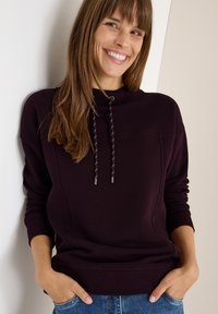 Felpa bordeaux con cappuccio, cordino decorativo e polsini a coste. Materiale morbido con dettagli sulle cuciture laterali. Abbinata a jeans blu.