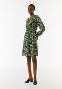 Grünes Leopardenmuster-Kleid mit langen Ärmeln, V-Ausschnitt und elastischem Taillenschnitt. Getragen mit schwarzen Absatzmules.