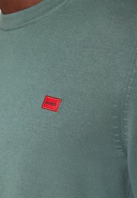 Ljusgrön stickad tröja med rund halsringning. Har en liten röd rektangulär patch med "HUGO"-logotyp. Slät textur och figurnära passform.