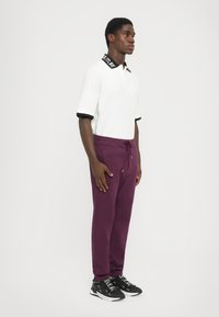 Camisa polo branca com colarinho e detalhes nas mangas em preto; calças de jogging roxas escuras com cordão; sapatilhas pretas com letras brancas.