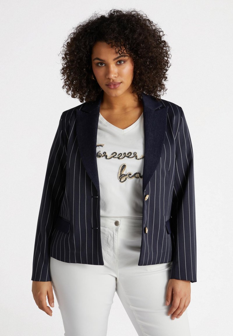 Blazer à rayures navy avec une coupe ajustée, doté de boutons et de revers dorés. Porté sur un t-shirt blanc orné et un pantalon blanc.