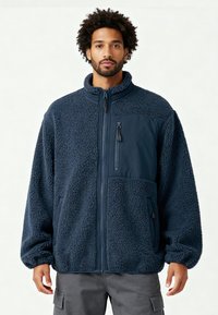 Veste polaire bleu marine avec un extérieur texturé et douillet ainsi que des panneaux en tissu lisse. Dotée d'un col montant et de poches zippées.