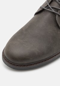 Chaussure habillée en cuir gris avec une texture lisse, un bout arrondi et des coutures marron contrastantes le long de la semelle. Les lacets renforcent un ajustement sécurisé.