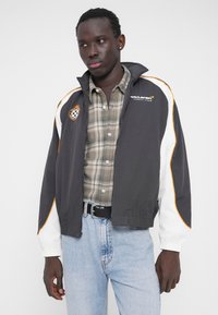 Μαύρο και άσπρο μπουφάν με πορτοκαλί λεπτομέρειες, με ψηλό γιακά και φερμουάρ. Φορεμένο πάνω από πουκάμισο με καρό μοτίβο και μπλε τζιν. Κανονική εφαρμογή.