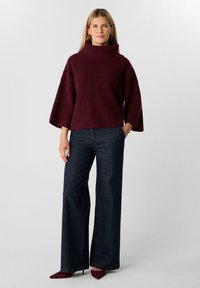 Woman wearing a burgundy turtleneck sweater with wide sleeves, dark wide-leg jeans, and burgundy pointed heels, standing against a plain background.