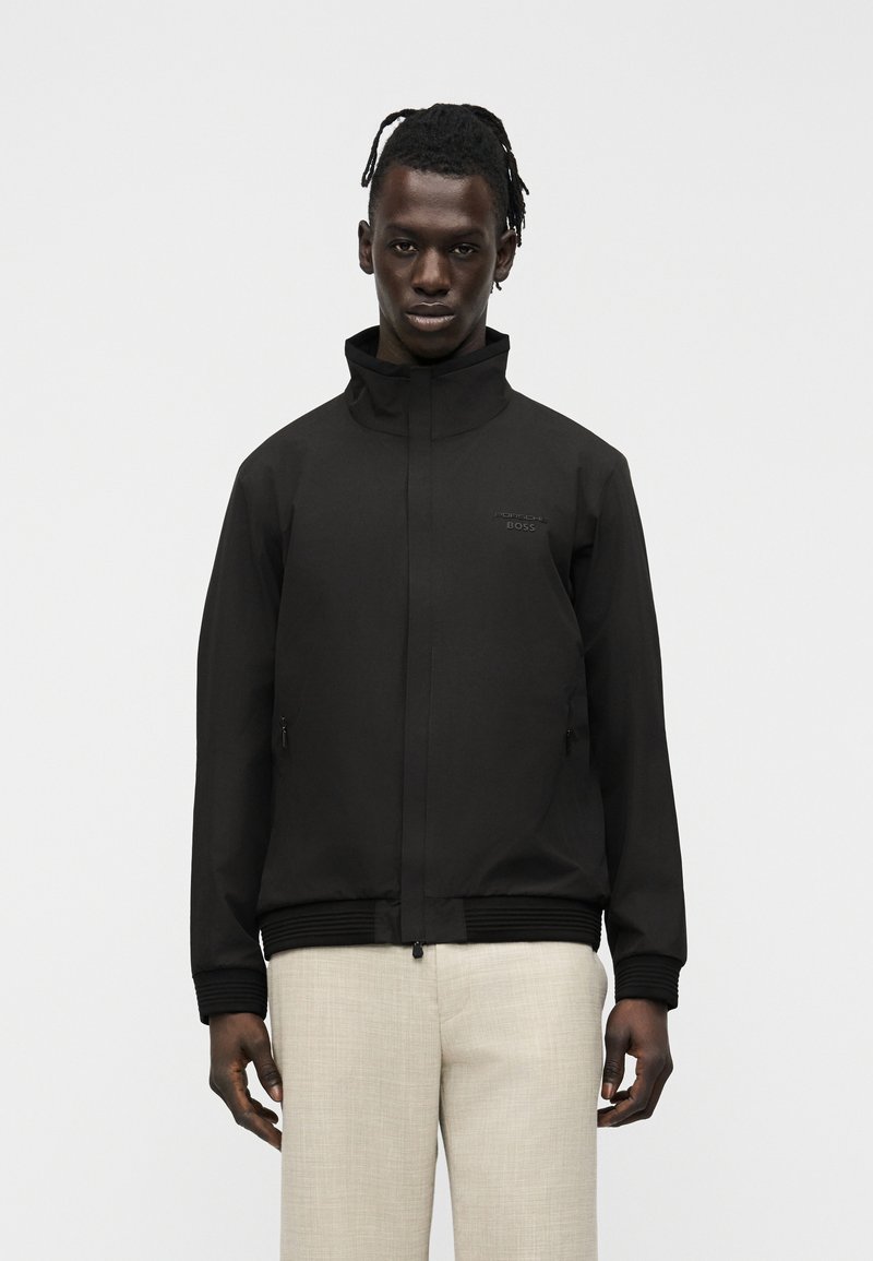 Άνδρας που φοράει μαύρο μπουφάν με φερμουάρ και ψηλό γιακά, καθώς και ανοιχτό μπεζ παντελόνι, στέκεται μπροστά σε απλό φωτεινό φόντο.