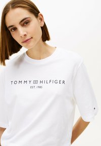 Camiseta blanca de algodón con el logo "TOMMY HILFIGER EST. 1985" impreso en azul oscuro. Manga corta y corte relajado. Diseño minimalista.