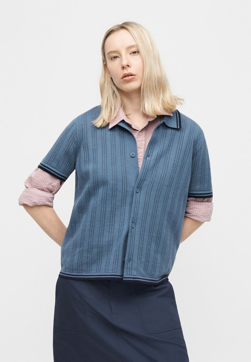 Donna bionda con capelli lisci indossa un cardigan di maglia blu a maniche corte sopra una camicia rosa a maniche lunghe arrotolata e una gonna scura.