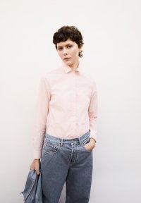 Lichtroze langemouwshirt met een kraag en knopen aan de voorkant, gecombineerd met blauwe high-waisted jeans. Het model houdt een spijkerjasje vast.