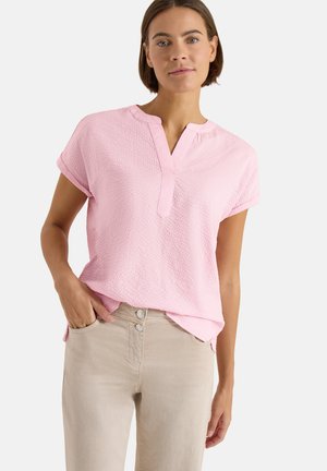 Kobieta ubrana w jasnoróżową, teksturowaną bluzkę z krótkim rękawem i rozciętym dekoltem oraz beżowe spodnie, stojąca z jedną ręką w kieszeni.