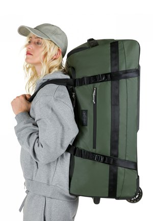 Borsa da viaggio verde con ruote, con cinghie nere, tasca laterale con zip e texture liscia, indossata sulla schiena da una persona con felpa grigia con cappuccio.