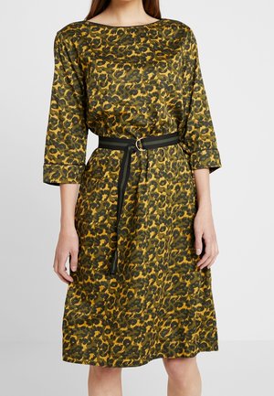 Femme portant une robe mi-longue à imprimé léopard vert et jaune, avec des manches trois-quarts et une ceinture noire avec une boucle ronde en argent.