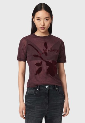 Jeune femme aux cheveux noirs raides portant un t-shirt transparent violet foncé à motif floral et un jean noir taille haute.