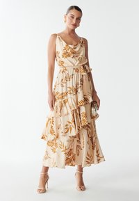 Abito lungo crema con stampa a foglia in ruggine, caratterizzato da un colletto drappeggiato, volant e vita legata. Tacchi a strappy argentati e una pochette.