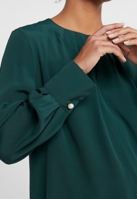 Blusa a maniche lunghe in tessuto verde scuro con una texture liscia, caratterizzata da un collo rotondo e un accento di perla sul polsino.