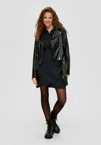 Schwarze Leder-Bikerjacke über einem dunklen Kleid mit asymmetrischem Saum, kombiniert mit schwarzen Strumpfhosen und schnürbaren Knöchelstiefeln.