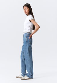 Νεαρή γυναίκα με μακριά σκούρα μαλλιά στέκεται πλαγίως, φοράει λευκό μπλουζάκι, φαρδιά μπλε τζιν και λευκά αθλητικά παπούτσια μπροστά σε ένα απλό φόντο.