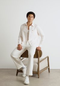 Camicetta button-up rosa chiaro, canotta bianca, pantaloni bianchi, sneakers bianchi con dettagli neri, seduto su una sedia di legno con seduta imbottita.