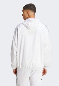 Veste à capuche blanche au fit ample, avec un ourlet élastiqué et de subtils accents noirs sur les côtés. Texture lisse et matériau léger.