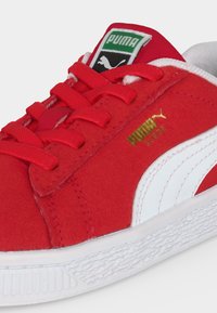 Baskets Puma en suède rouge avec bande latérale blanche, lacets rouges, semelle blanche texturée et logo Puma vert sur la languette.
