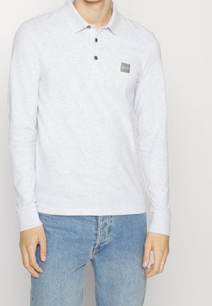 Mann trägt ein hellgraues Langarm-Polohemd mit drei Knöpfen und kleinem "BOSS"-Logo-Patch, kombiniert mit blauen Jeans.