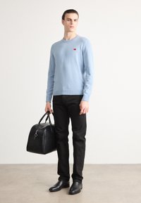 Hellblauer Strickpullover mit kleinem rotem Logo, schwarze Jeans und schwarze Ledertasche. Geschmeidige Textur, Rundhalsausschnitt und taillierte Silhouette.