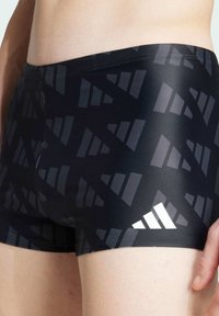 Čierne plavecké šortky s geometrickým vzorom v rôznych odtieňoch šedej. Majú elastický pás a výrazný biely logotyp.