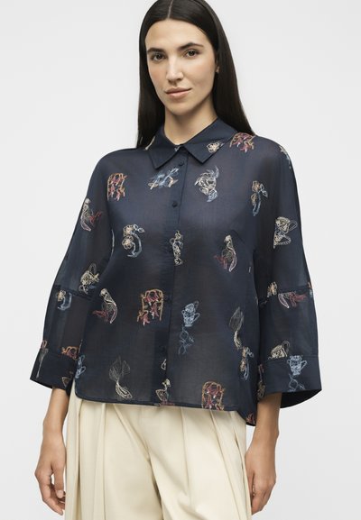 Femme aux longs cheveux foncés porte un chemisier ample bleu marine à boutons avec une broderie abstraite colorée et un pantalon plissé beige clair.
