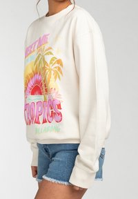 Žena v bílém mikině s barevným potiskem tropického západu slunce a palmy, doplněná o roztrhané džínové šortky.