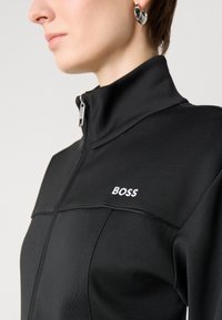 Black athletic jacket featuring a high collar, front zip, and white "BOSS" logo. Smooth fabric with structured seams for a fitted design.