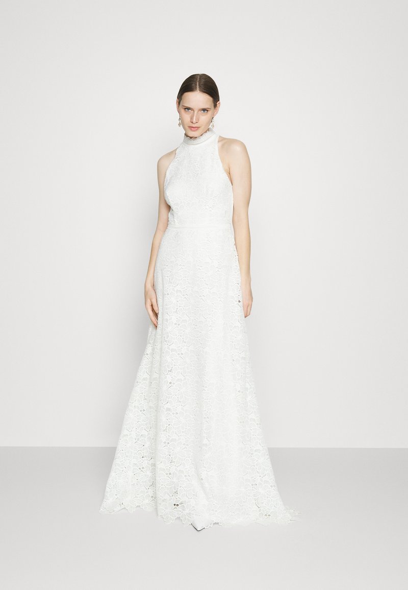 Mujer vestida con un vestido de encaje blanco sin mangas y de cuello alto, con un patrón floral, de pie contra un fondo claro liso.