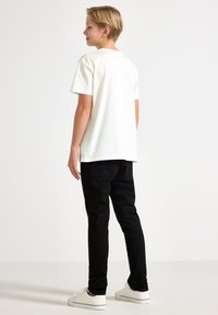 Bílý bavlněný tričko s krátkým rukávem a kulatým výstřihem, v kombinaci s černými denimovými kalhotami a bílými teniskami, viděno zezadu.