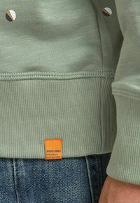 Miętowo-zielona bluza dresowa o prążkowanej fakturze, z małymi, wielokolorowymi okrągłymi wzorami oraz pomarańczową metką u dołu.