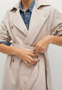 Beige trenchcoat met een ceintuur in de taille, dubbelzijdige knoopsluiting en grote revers. Zacht materiaal met opgerolde mouwen.