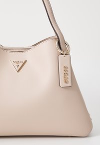 Beige käsilaukku, valmistettu sileästä synteettisestä materiaalista, jossa on kolmiomainen kultainen logo, kultainen rengaskoriste ja pieni silmukka, johon on kiinnitetty merkitty tunniste.