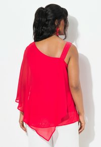 Top in chiffon rosso con design asimmetrico, spalla scoperta, spalline decorate e silhouette fluida, indossato con pantaloni bianchi.