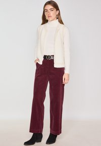 Gilet senza maniche color avorio con tessuto testurizzato sopra un dolcevita bianco aderente; pantaloni a gamba larga in velluto a coste borgogna; cintura nera con fibbia circolare.