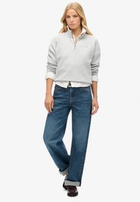 Grijze sweatshirt met een ritskraag, gedragen over een wit shirt, gecombineerd met loszittende blauwe jeans en zwarte schoenen met opgerolde broekspijpen.