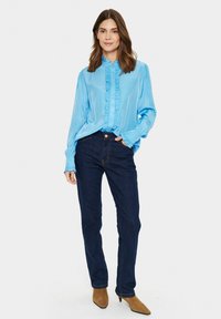 Blusa di raso azzurro chiaro con ruches sul davanti e ai polsini, abbinata a jeans di denim blu scuro e stivaletti marroni a punta.