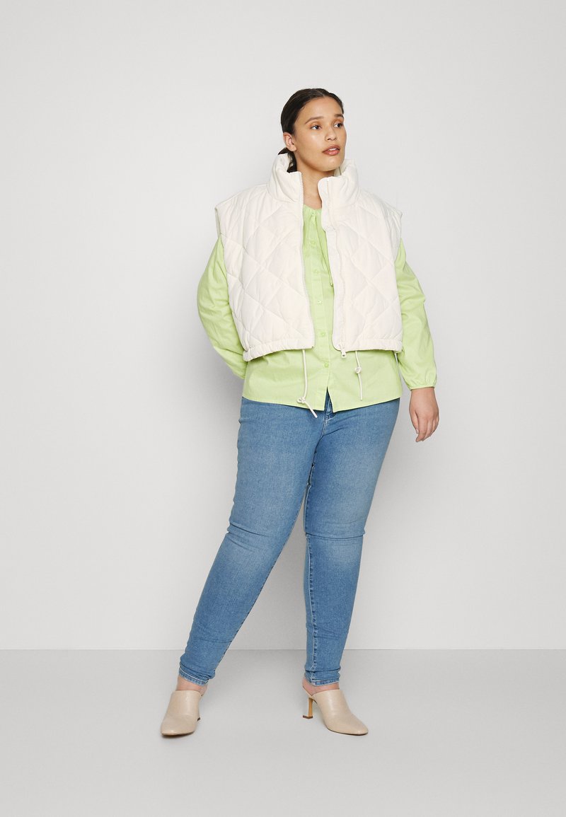 Kurtka w kolorze jasnego beżu, pikowana, z wysokim kołnierzem i elastycznym dołem. W zestawie z jasnozieloną koszulą zapinaną na guziki oraz niebieskimi jeansami o wąskim kroju.