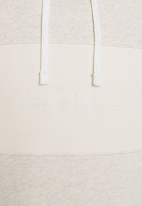 Hoodie beige clair avec une partie supérieure texturée et une partie inférieure lisse, présentant un logo blanc brodé et un cordon de serrage.