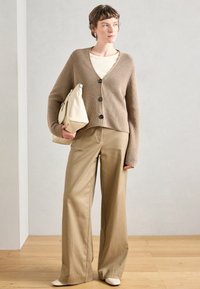 Cardigan bege com textura canelada e botões pretos, combinado com calças largas de tom castanho claro e uma bolsa creme. Textura suave, ajuste descontraído.