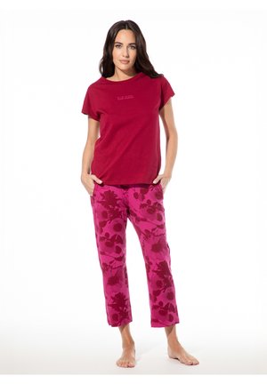 Frau steht barfuß, trägt ein rotes Kurzarmshirt und rosa Hose mit Blumenmuster, die Hände in den Taschen auf weißem Hintergrund.