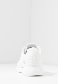 Valkoinen liikuntakenkä, jossa on teksturoitu verkkopinta, pehmustettu kaulus ja kumipohja. Sisältää hillityn logon kantapäällä ja virtaviivaisen muotoilun.