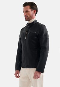 Zwarte neplederen jas met hoge kraag, ritszak op de borst en geribbelde schouderaccenten. Glad textuur en op maat gemaakte pasvorm.