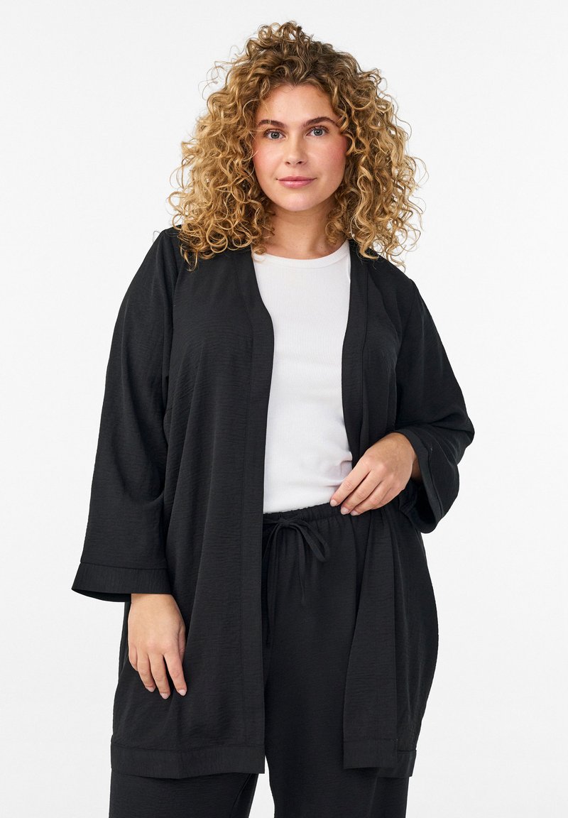 Mujer con cabello rizado y rubio lleva un cárdigan negro suelto abierto sobre una blusa blanca y pantalones negros a juego con cordón, posando contra un fondo blanco.