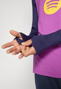 Camisola atlética de mangas compridas com um corpo roxo e mangas azul-marinho, com buracos para os polegares, acentuada por um grande logótipo amarelo. Tecido suave e elástico.