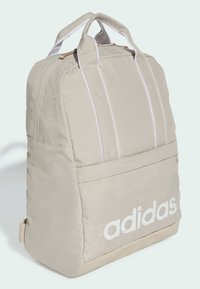 Mochila de nylon beige con acentos blancos, dos asas y bolsillo frontal con un gran logo blanco. Diseño acolchado para mayor comodidad.